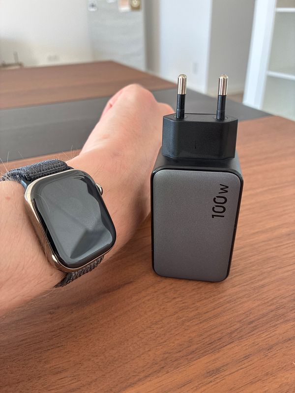 A hand wearing a smartwatch rests on a wooden table beside a compact 100W UGREEN charger with a European plug.