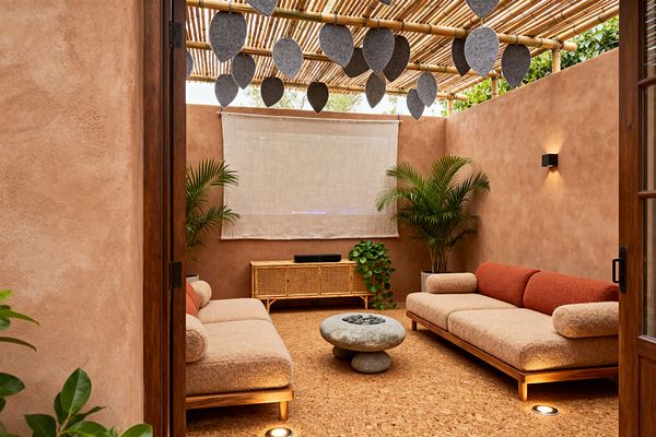 Cozy media room with terracotta plaster walls and a bamboo pergola overhead, hung with gray felt leaf baffles, where two low wood-framed sofas in beige bouclé with rust cushions face a rounded stone coffee table. A rattan-cane console with a soundbar sits beneath a linen projection screen, flanked by potted palms and trailing greenery, while wall sconces and in-floor uplights provide warm, soft illumination.