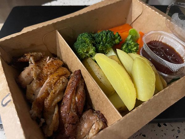 A takeaway box containing grilled chicken, vegetables, and dipping sauce.