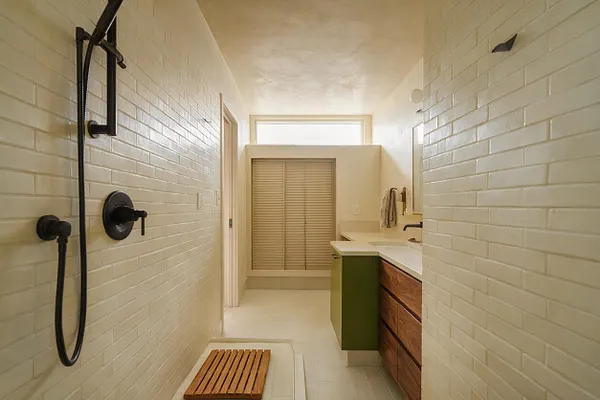 A narrow master bathroom with white subway tile walls, matte black shower fixtures, and a low shower floor with a wooden slatted mat. At the far end, a vanity with walnut drawers, an olive green side panel, and a light countertop sits beneath a mirror, with a small window and louvered closet door beyond.