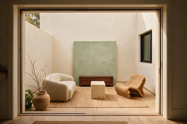 A small enclosed backyard courtyard seen through a large sliding opening, furnished with a cream lounge chair, a woven sculptural chair, a low wood bench, and a pale stone cube table on a natural jute rug. Soft white stucco walls, a muted green wall panel, and simple pottery with bare branches create a calm organic modern atmosphere.