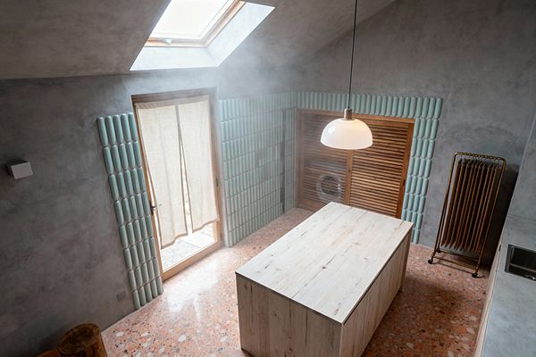 A bright laundry room features a central light wood island beneath a skylight. Seafoam green, rounded ceramic tiles line the lower walls and frame a wood-framed glass door with a linen curtain and a louvered timber cabinet concealing the washer. The floor is warm pink terrazzo and the walls are soft gray plaster. A white dome pendant, brass drying rack, and a compact stainless sink complete the airy coastal look.