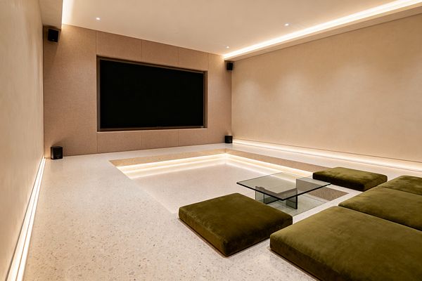 A minimalist media room features a sunken seating pit in pale terrazzo with steps lit by linear LED strips. Olive green velvet floor cushions and a low clear glass table face a large black screen recessed in beige fabric acoustic panels, with small black speakers at the corners. Smooth warm-toned plaster walls and concealed cove lighting at floor and ceiling create a softly lit, uncluttered viewing space.