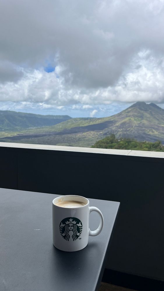 A Starbucks coffee mug sits on a table with a scenic view of mountains and clouds in the background.