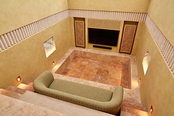 A sunken media room features travertine steps and floor forming a central pit, surrounded by ochre plaster walls with a fluted band near the ceiling. A large flat-screen with a black soundbar is framed by wood lattice panels; two backlit marble niches punctuate the walls, recessed step lights wash the stone in warm glow, and a long sage-green upholstered sofa with nailhead trim sits on the upper tier.