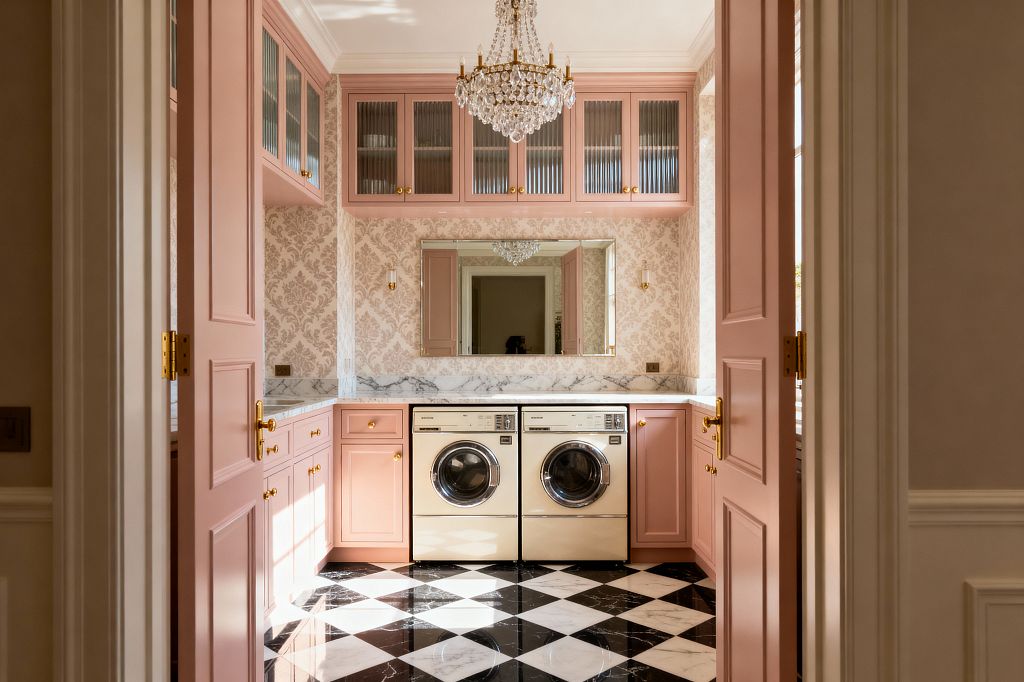 A vintage-glam laundry room viewed through pink double doors features blush shaker cabinets with brass knobs, a white marble countertop and short backsplash, and a pair of cream front-loading machines with chrome trim. Upper cabinets with ribbed glass flank a wide mirror over damask wallpaper, while a crystal chandelier and two small brass sconces add sparkle. The floor is a polished black-and-white checkerboard marble laid on the diagonal, brightened by natural light and classic crown molding.
