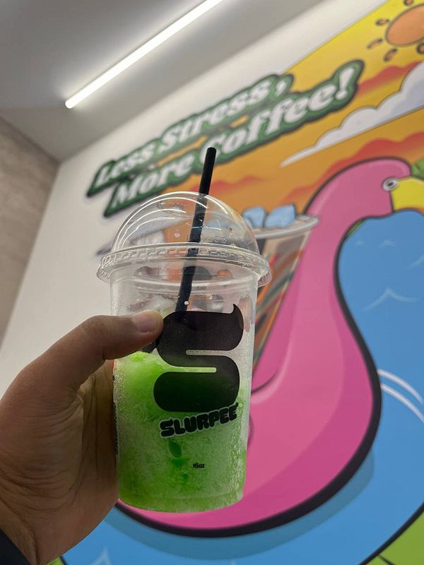 A hand holds a green Slurpee in front of a colorful mural.