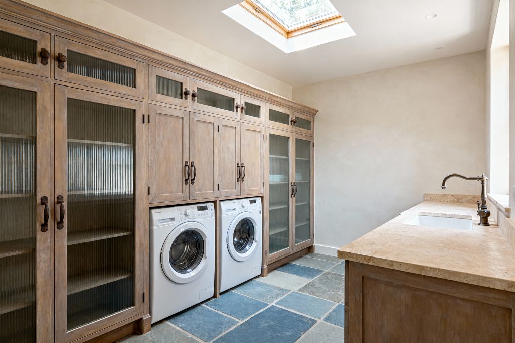 A bright laundry room features a full wall of weathered wood cabinets with reeded-glass doors and dark bronze pulls, with two white front-loading machines set below. Opposite a long beige stone countertop with a rectangular white sink and bronze gooseneck faucet sits under a window, while large blue-gray slate tiles cover the floor and off-white plaster walls and a ceiling skylight fill the space with natural light.