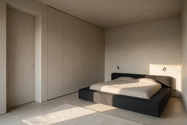A minimalist master bedroom with a low black platform bed dressed in light bedding, set against soft beige walls and built-in closet panels. Natural sunlight creates a warm rectangular patch on the wall and floor, highlighting the calm, uncluttered space.