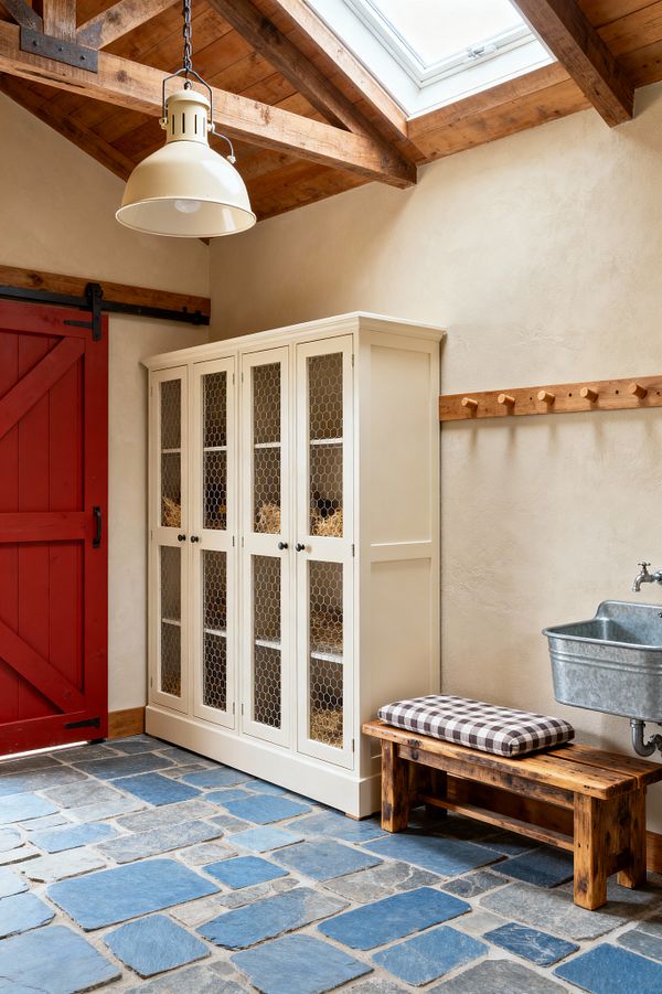A farmhouse-style mudroom with a barn-red sliding door on black hardware, a cream cabinet with chicken-wire panel doors, and a rustic wooden bench topped with a brown-and-cream gingham cushion. The space features blue-gray bluestone flooring, textured beige plaster walls with a wood peg rail, a wall-mounted galvanized utility sink, exposed wood ceiling beams with a skylight, and a cream enamel pendant light.