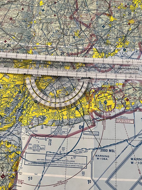 A detailed aviation plotter tool with navigational markings, scales, degree measurements, and a pencil.