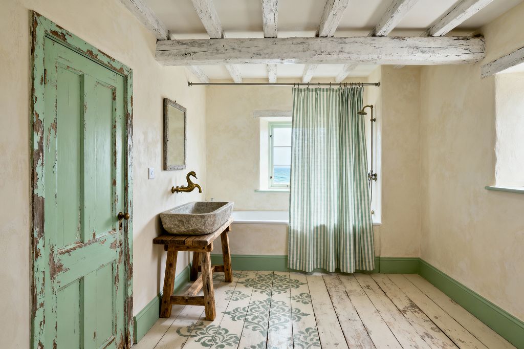 An airy shabby chic bathroom features a distressed mint-green door, matching skirting and window trim, and whitewashed ceiling beams over warm cream plaster walls. A carved stone trough sink sits on a rustic trestle table beneath a weathered wood mirror, with a brass swan-neck wall spout. Lime-washed plank floors carry sage stenciling, while a green-and-white striped cotton curtain softens a tub with exposed brass shower beside a small sea-facing window.