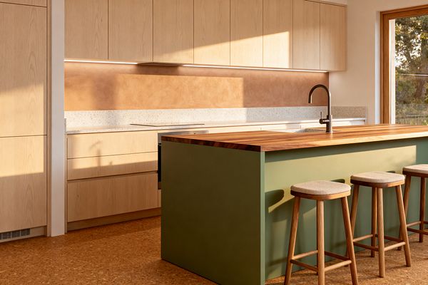A sunlit Scandinavian kitchen shows flat-front light oak cabinets, a speckled light terrazzo countertop, and a terracotta-toned plaster backsplash illuminated by a slim under-cabinet light. A sage green island with a thick butcher-block wood top holds a brushed stainless faucet and undermount sink, with three wooden stools upholstered in beige fabric. Natural cork flooring and a wood-framed window complete the calm, minimal space featuring clean lines and warm materials.