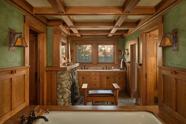 A Craftsman-style master bathroom with warm wood paneling, exposed coffered ceiling beams, and green walls. The room features a stone-faced fireplace, a built-in wood vanity with dual sinks and a large window, wall sconces, and a clawfoot-style tub in the foreground.