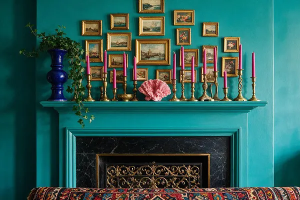 A vibrant teal fireplace mantel is styled with a blue vase and trailing greenery, a row of brass candlesticks with pink candles, and a pink shell-like decorative object. Above it, a cluster of small gilt-framed landscape and floral paintings creates a dense gallery wall, with a black marble firebox and ornate brass screen below.