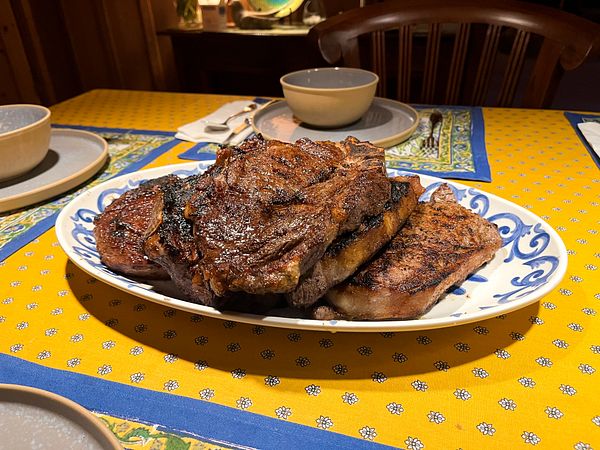 A plate filled with several grilled steaks placed on a yellow floral tablecloth with blue and green patterned napkins and tableware around it.