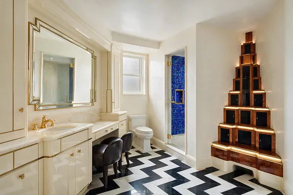 A bright Art Deco master bathroom with glossy cream cabinetry, a large framed mirror over a marble vanity, and a black-and-white chevron floor. The room includes a glass shower with vivid blue mosaic tile and a dramatic illuminated stepped wall shelf in warm wood and black finishes.