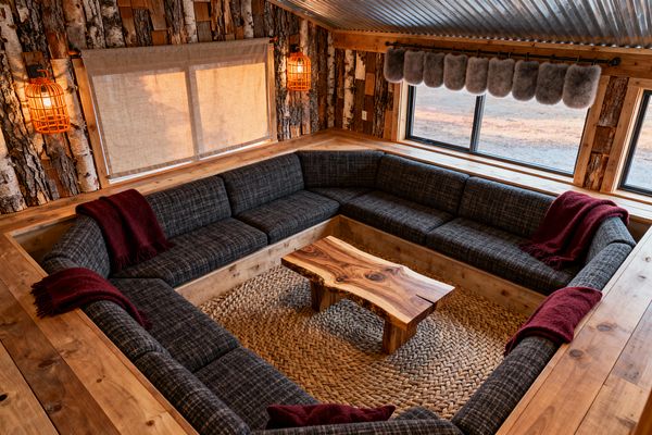 A cozy, sunken media room forms a square conversation pit built from knotty pine with charcoal tweed cushions and burgundy wool throws. A live-edge walnut coffee table sits on a braided jute rug. Walls mix birch-bark cladding and reclaimed wood, illuminated by warm caged lantern sconces. A corrugated metal ceiling adds texture. One window has a linen roll shade, while another features a gray shearling valance on a black rod overlooking open land.