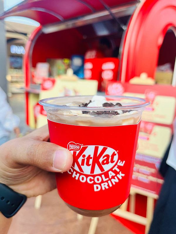A hand holds a cup of KitKat chocolate drink in front of a vibrant red kiosk.