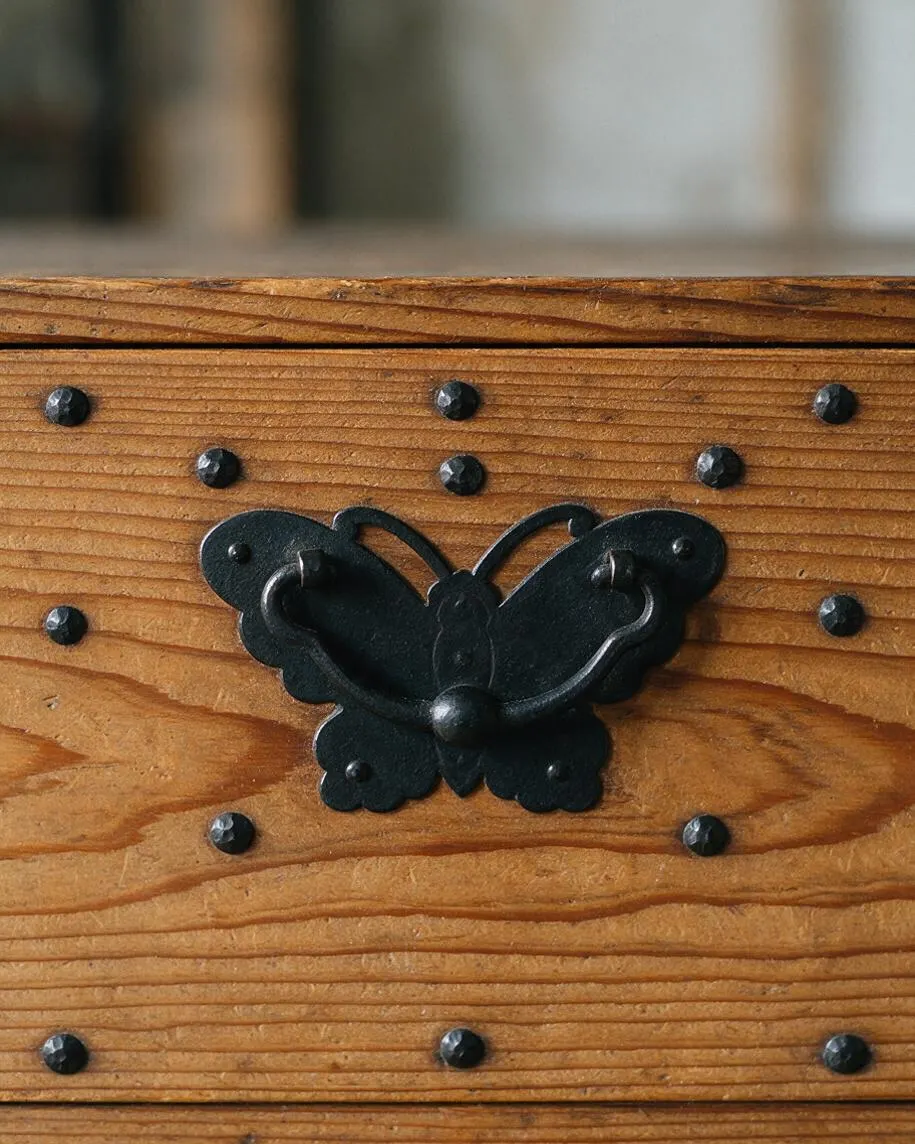 A close-up of a wooden chest or cabinet front made of warm, textured cedar planks with visible grain and dark nail heads. Centered on the panel is a decorative black iron butterfly-shaped handle or latch, creating a traditional Japanese-inspired Zen detail.