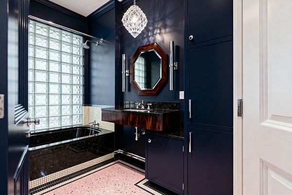 An Art Deco bathroom with deep navy walls and cabinetry, a floating wood-veneer vanity topped with black granite, and an octagonal wood-framed mirror flanked by tall chrome sconces beneath a faceted crystal pendant. A tub-shower with black stone surround and cream mosaic trim sits before a large glass-block window, while pink terrazzo flooring with a black-and-white border and polished chrome fixtures complete the space.