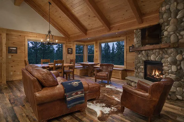 A rustic studio apartment with a vaulted wood plank ceiling, knotty pine wall paneling, and large picture windows overlooking trees. The room features a stone fireplace with a mounted TV, a leather sofa and armchairs, a wooden dining table, and warm ambient lighting.