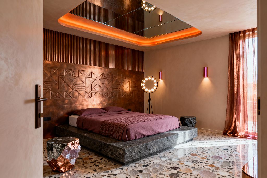 A Cosmic Chic bedroom features a burnished copper headboard wall with vertical slats and embossed geometric patterns behind a low, dark-stone platform bed dressed in mauve linen. The room includes terrazzo flooring, a faceted geode side table with copper sheen, rose sheer curtains, magenta metal wall sconces, a halo floor lamp, and a mirrored ceiling recess with orange cove lighting that warms the blush plaster walls.
