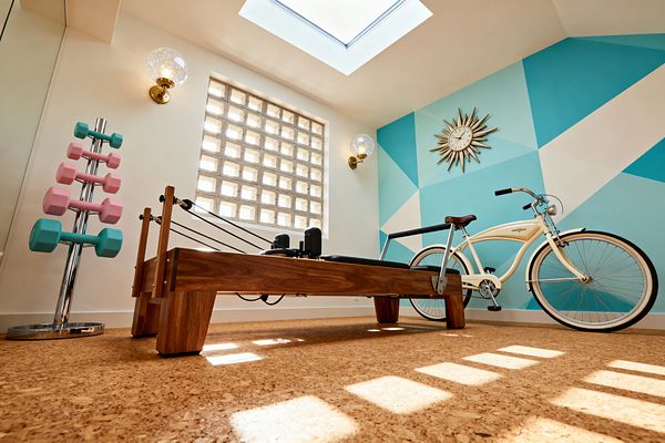 Bright home gym with a wooden Pilates reformer sits on cork flooring beneath a large skylight. A teal and aqua geometric mural with a brass sunburst clock spans one wall, while brass-and-glass globe sconces flank a square glass-block window. A chrome dumbbell tree holds pastel pink and turquoise weights, and a cream cruiser bicycle rests nearby, creating a clean, retro atmosphere with natural wood, warm light, and crisp lines.