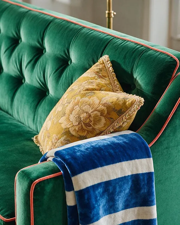 A close-up of a deep emerald green velvet sofa with tufted upholstery and coral piping along the edges. A gold floral cushion and a blue-and-white striped throw blanket add layered texture and rich color, creating a luxurious maximalist look.