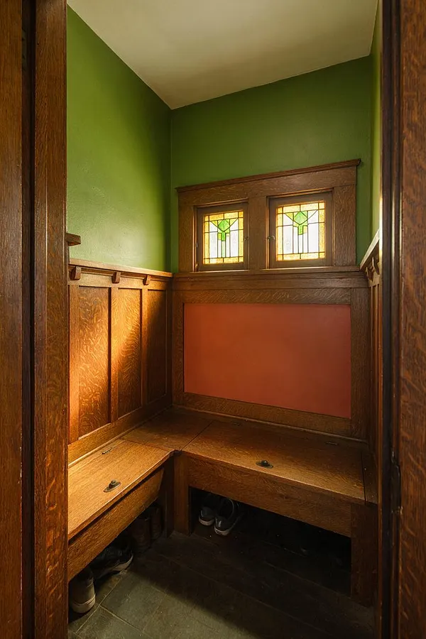 A compact Craftsman-style entryway with rich wood paneling, built-in corner benches, and green painted walls. Two small stained-glass windows above a coral-toned wainscot panel bring in warm light.