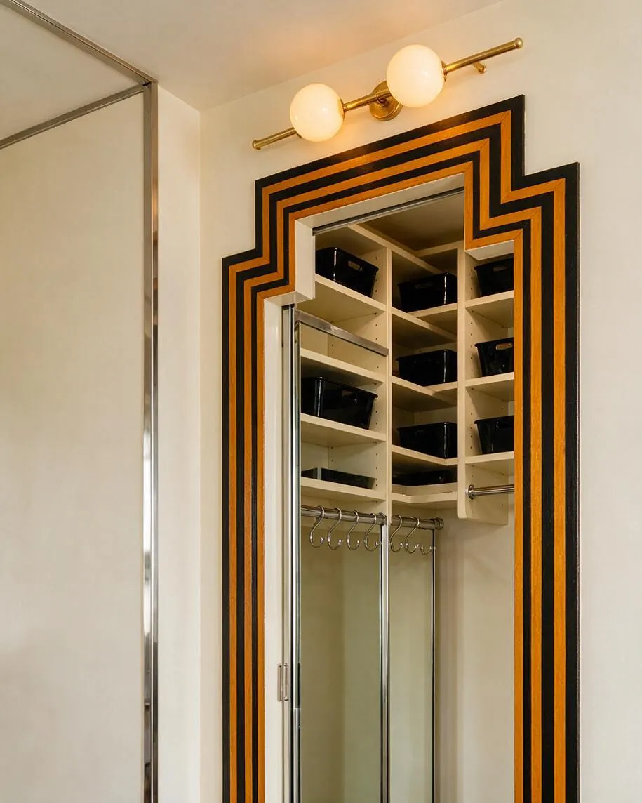 A compact closet features cream shelving, chrome hanging rods, and black storage bins, framed by a bold stepped Art Deco mirror trim in black and gold. Above it, a brass sconce with two round frosted globes casts a warm glow, while a mirrored sliding door reflects the clean, elegant interior.