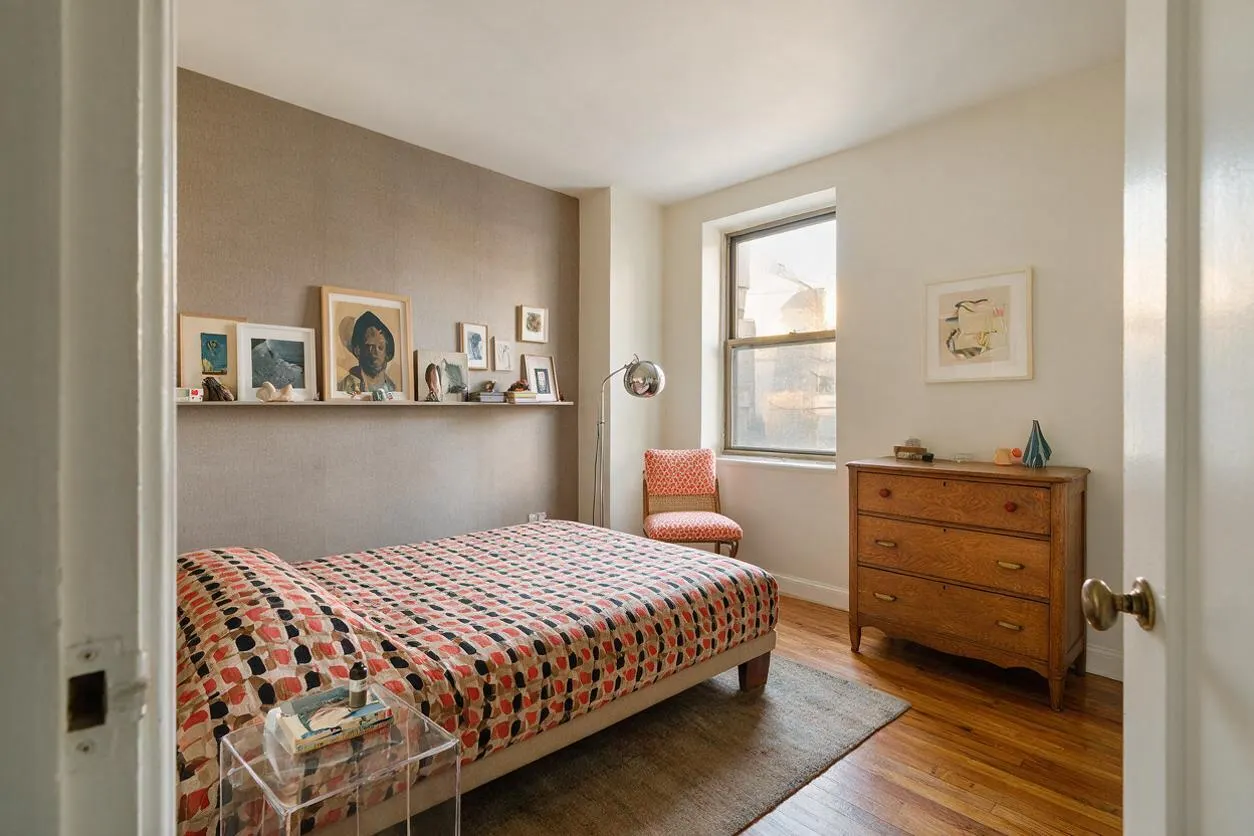 A compact eclectic bedroom with a low bed covered in a patterned quilt, a gallery shelf of framed art along one wall, and a warm wooden dresser by a sunlit window. A pink patterned chair, chrome floor lamp, clear side table, and wood floors add a collected, lived-in feel.