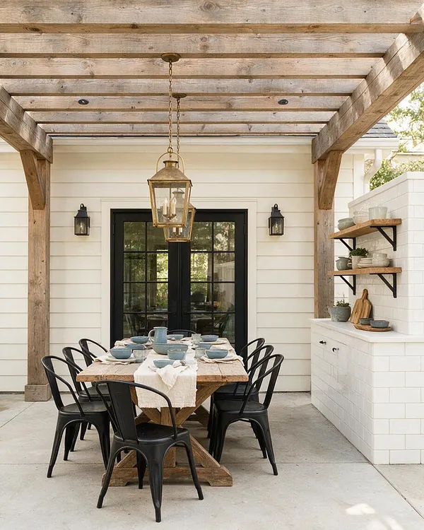 A covered backyard dining area in farmhouse style with a weathered wood pergola overhead, a hanging lantern light, and black French doors on a white shiplap wall. A rustic wood table is set with muted blue dishes and surrounded by black metal chairs, while white brick shelves and a built-in counter hold simple decor on the right.