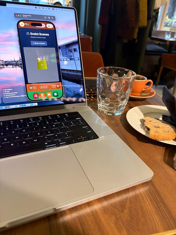 A laptop displaying the Godot game engine interface is set on a wooden table alongside a glass of water and a cookie.