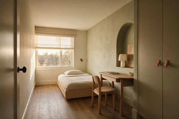 A narrow kids' room with a low wood bed, pale bedding, and a small wooden desk and chair along one wall. Soft beige walls, an arched niche with a lamp, and a large window with a sheer shade create a calm, natural atmosphere.