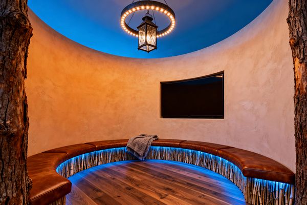 A circular media room with warm plaster walls features a curved saddle-brown leather banquette around the perimeter, its base wrapped in vertical bamboo reeds illuminated by blue LED strips. A flat-screen TV is set into the wall. Overhead, a black iron ring chandelier with a central lantern hangs beneath a blue-lit dome ceiling. Wide-plank walnut floors and rough tree-trunk columns complete the rustic setting, with a gray throw on the seat.
