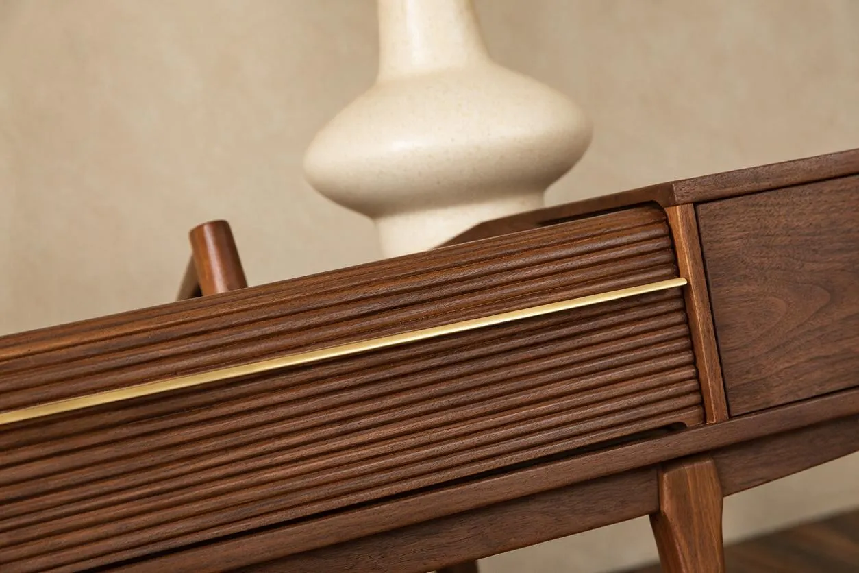 A close-up of a mid-century modern wooden cabinet or credenza with richly grained walnut, horizontal reeded drawer fronts, and a slim brass accent line. Tapered wooden legs and a soft sculptural vase in the background reinforce the warm, streamlined style.