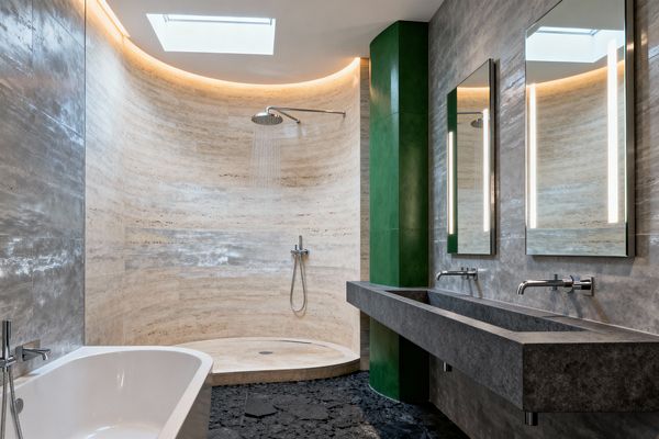 A modern bathroom features a curved walk‑in shower lined in beige travertine with a round rain showerhead, beside a freestanding white bathtub. Along the right wall, a long dark gray concrete trough vanity holds two wall‑mounted chrome faucets below twin mirrors with vertical LED strips, while a green column, a dark irregular stone floor, and a skylight with cove lighting complete the space.