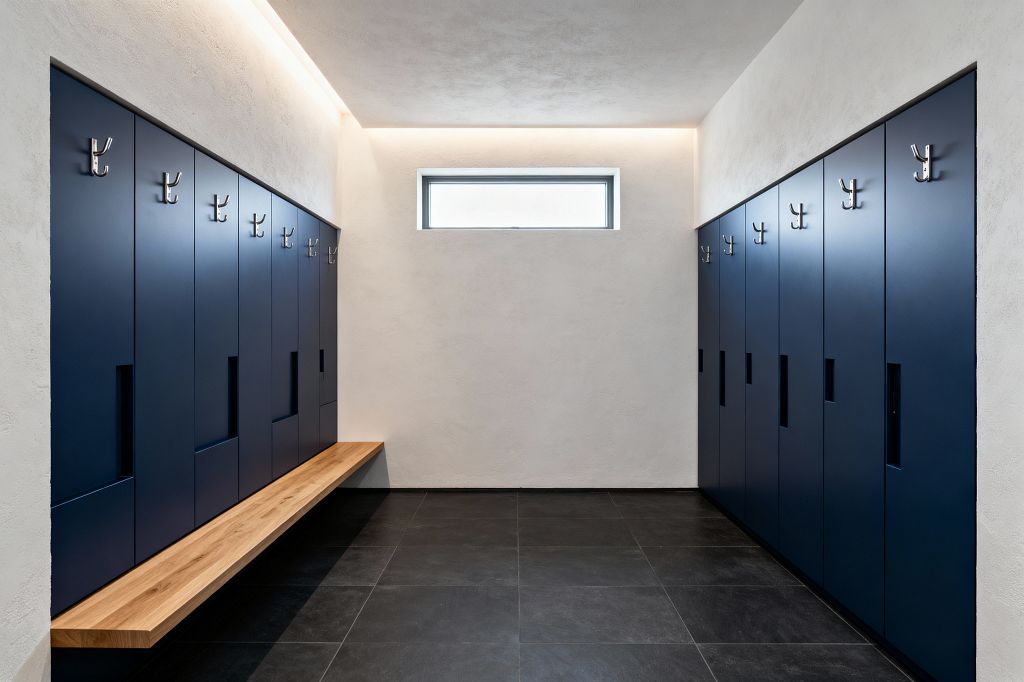 A minimalist mudroom features two rows of matte navy built-in lockers with recessed pulls and brushed metal double hooks, forming a symmetrical corridor. A floating oak bench runs along the left side above large charcoal porcelain tiles, while off-white plaster walls, a narrow horizontal clerestory window, and soft perimeter cove lighting keep the space bright, durable, and uncluttered.