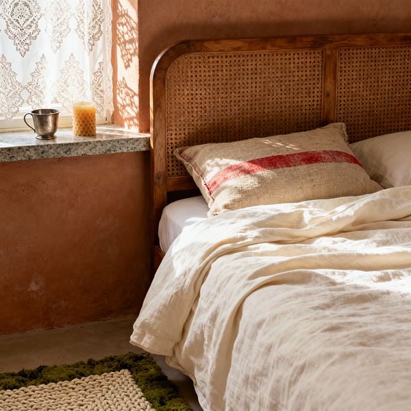 A cozy farmhouse bedroom corner features a wood-framed cane headboard against warm terracotta plaster walls, dressed with ivory linen bedding and a rustic grain-sack pillow with a red stripe. Sunlight filters through lace curtains, casting patterns on a gray granite windowsill holding a beeswax candle and small metal cup; at the bed’s edge sits a chunky knit natural-fiber rug with a moss-green border.