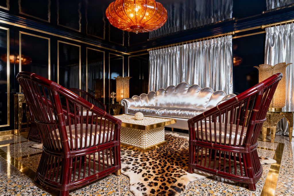A glamorous living room with high-gloss black lacquered walls trimmed in gold, featuring a scalloped silver satin sofa and two oxblood rattan armchairs around a pearl-studded brass coffee table. An amber glass pendant, shimmering silver curtains, a leopard-print hide rug, terrazzo flooring with brass inlays, and ornate gold lamps create a richly reflective, dramatic setting.