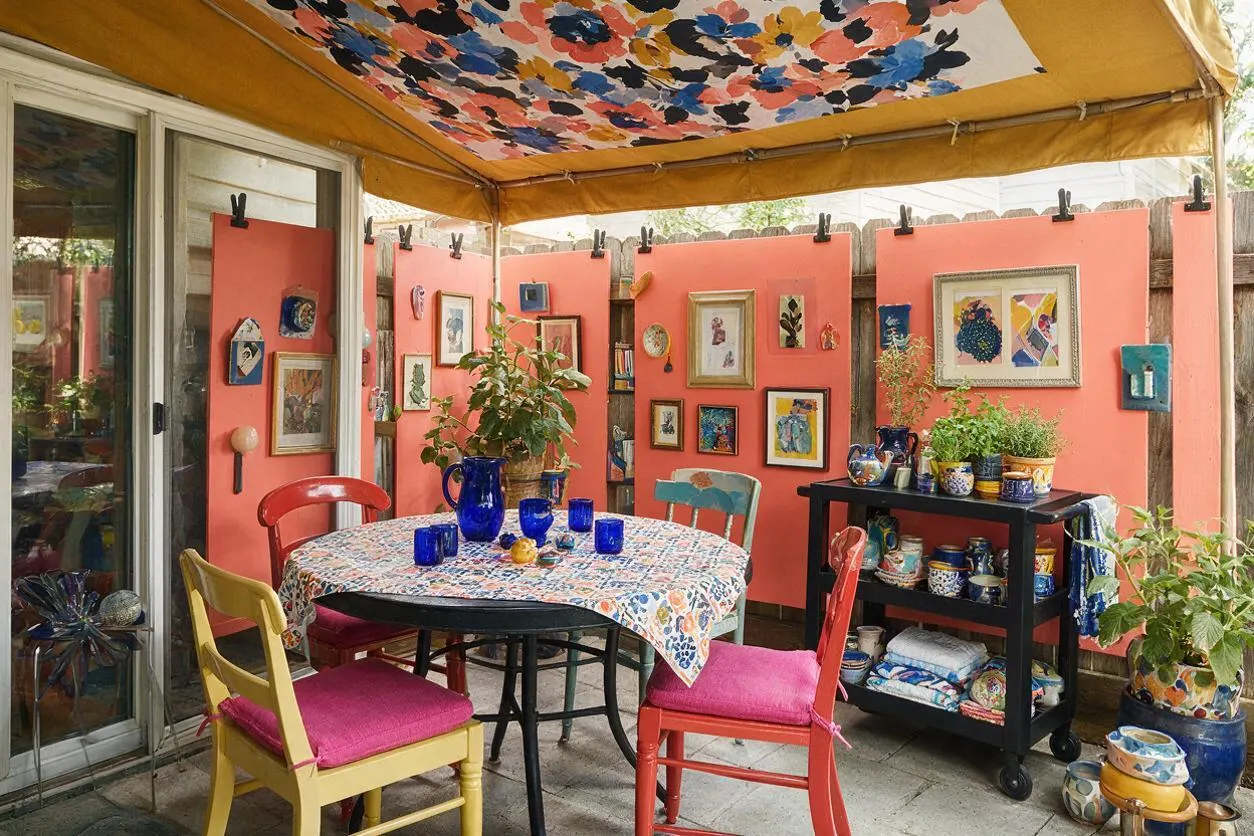 A covered backyard patio filled with coral-painted walls, a floral canopy overhead, and a round table set with blue glassware. Mismatched painted chairs, framed artwork, potted plants, and shelves of ceramics create a lively maximalist outdoor dining space.
