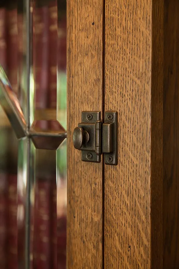 A close-up view of a Craftsman-style wood cabinet door showing warm oak grain and a dark bronze hinge with visible screws. The adjacent leaded glass panel has faceted geometric panes that reflect soft light and add a traditional handcrafted look.