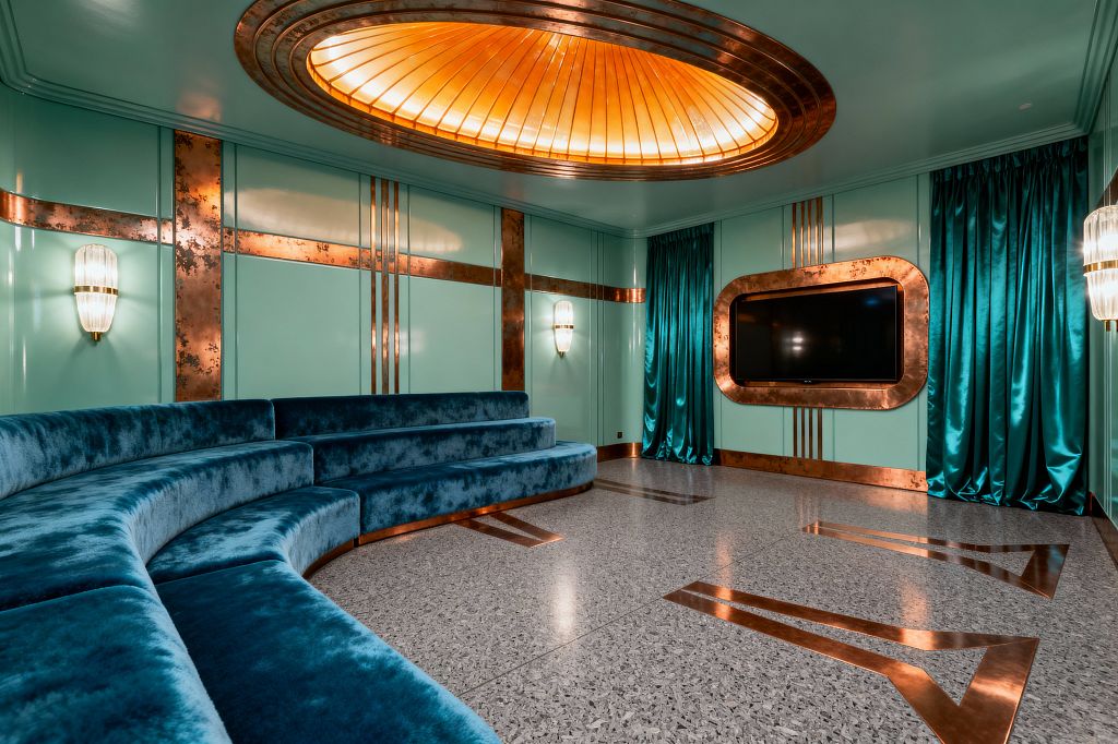 A glamorous Art Deco media room with glossy mint wall panels trimmed in patinated copper. A curved, tiered banquette in deep teal velvet wraps the space, facing a TV set into a rounded copper frame. The terrazzo floor is inlaid with copper chevrons. Overhead, an oval backlit amber glass dome is ringed in copper. Teal satin curtains and ribbed glass wall sconces complete the room’s rich, streamlined look.