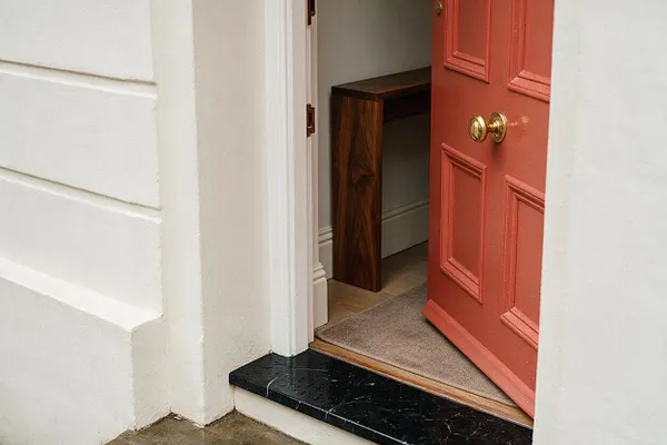 A close-up view of a home entry showing a coral-red paneled door with brass hardware set into a white exterior wall. A black marble threshold, beige doormat, and a glimpse of a warm wood table inside add texture and contrast.