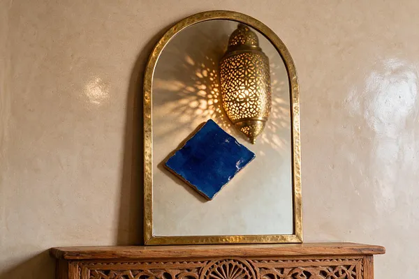 A brass-framed arched mirror hangs on a textured beige wall, reflecting a pierced Moroccan lantern that casts intricate light patterns. A cobalt blue diamond-shaped tile is mounted on the wall below the reflection, and the top edge of a carved wooden cabinet appears at the bottom of the frame.