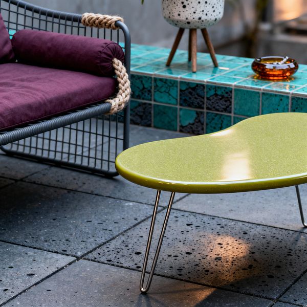A mid-century patio vignette with an olive green, kidney-shaped coffee table on hairpin legs sitting on speckled charcoal concrete pavers. To the left is a lounge chair with a charcoal metal rod frame, deep plum seat and bolster cushions, and thick natural rope wrapped around the arm. Behind, a low ledge is faced in teal glazed tiles with dark variegation. On the ledge sit a speckled white ceramic planter on a small wooden tripod and a small amber glass dish.