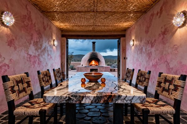 An open-air Southwestern dining room features mottled pink plaster walls, a woven reed ceiling, and a charcoal cobblestone floor centered by a long, polished marble slab table with bold veining. Eight dark-wood chairs wrapped in thick jute rope flank the table, amber glass votives and a clay bowl sit on top, faceted wall sconces sparkle, and open doors reveal a domed kiva-style wood oven glowing against a rugged desert backdrop.