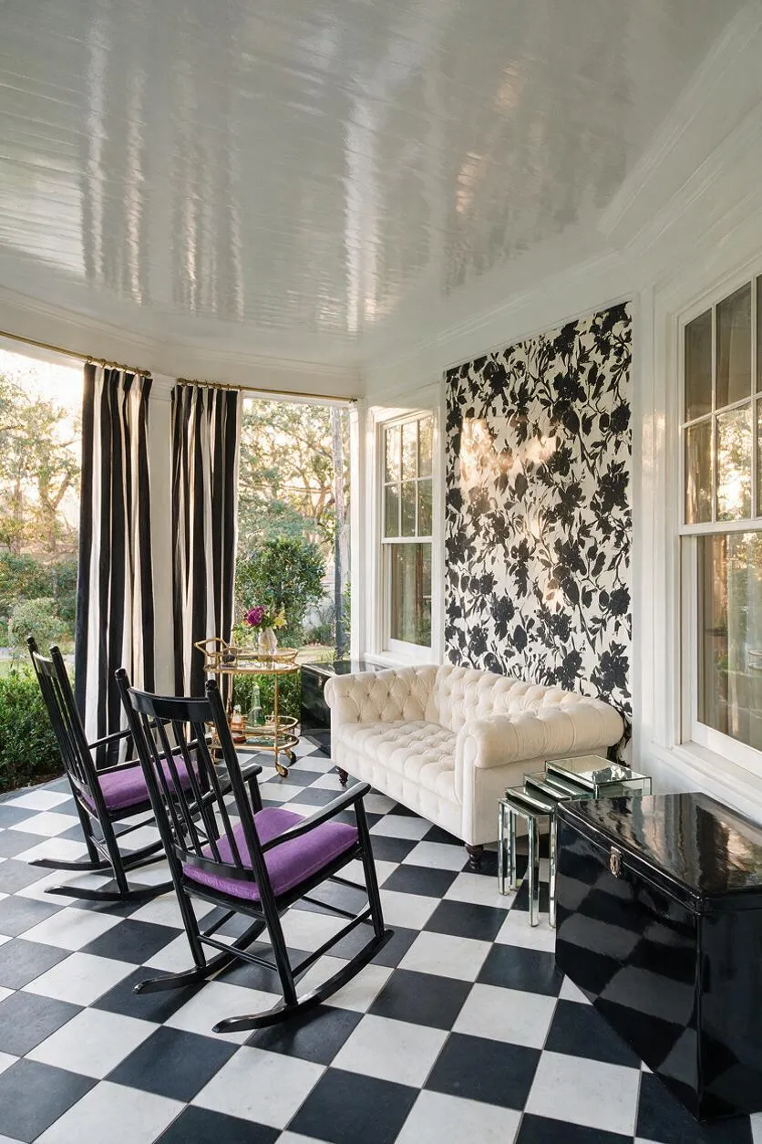 A stylish front porch with a glossy white ceiling, black-and-white checkerboard floor, and large windows framing garden views. Black rocking chairs with purple cushions sit near a tufted white sofa, mirrored side tables, and bold black-and-white floral wall covering, creating a glamorous Hollywood Regency look.