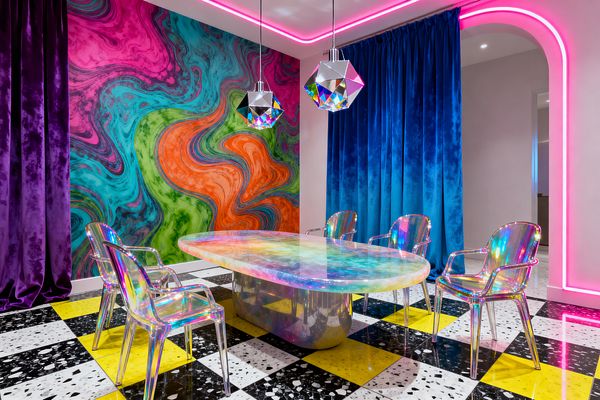 Dining room with a swirling marbled mural in magenta, teal, lime, and orange behind an oval resin table with a pearlescent surface and mirrored pedestal. Four translucent iridescent armchairs sit on a black-and-white speckled terrazzo and yellow checkerboard floor, framed by plush purple and cobalt velvet curtains; a pink LED strip outlines the ceiling and arched doorway, while two faceted chrome-iridescent pendant lights hang above the table.
